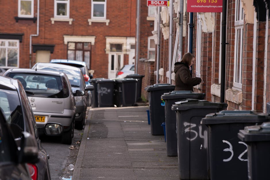 The image displays a narrow residential street lined with red brick terraced houses, each with white-framed windows and traditional stone lintels. On the right side of the street, a woman is standing outdoors near the front of one of the houses, positioned beside a row of large, dark grey wheelie bins with black lids, used for rubbish collection services. The bins are placed directly on the pavement, which is made of concrete slabs, some showing minor wear and dirt. Several of these waste containers are positioned close together, with some showing white graffiti markings or tags. The woman appears to be handling or sealing something inside the bin, dressed in a dark coat with a hood. To the left of the scene, parked cars are lined up along the street, including a small silver hatchback and a larger vehicle beyond it, their surfaces reflecting ambient daylight. The overall environment suggests an everyday scene of household waste management, associated with private or independent rubbish collection services, set amidst typical urban residential surroundings.