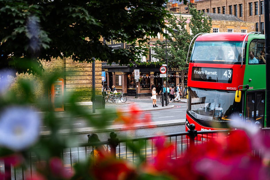 A double-decker bus, predominantly green and red with a black front, is seen on a busy street, displaying the route number 43 and the destination 'Friern Barnet' on a black digital sign. The bus is positioned toward the right side of the image, with its front and side windows visible, revealing some reflections of the surrounding environment. In the background, there are pedestrians walking along the sidewalk and waiting at the crosswalk, some with bicycles parked nearby, and a shopfront with a sign that reads 'The York.' The surrounding buildings are made of brick with a traditional appearance, featuring multiple windows and some decorative architectural elements. The street scene is framed partially by leafy green trees and out-of-focus flowers in the foreground, adding color and depth to the image. The overall atmosphere suggests a typical urban setting with active public transportation, pedestrian activity, and a mix of historical and modern urban features, aligning with contexts where private or independent waste collection might be relevant in the broader city environment.