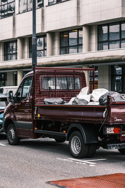 A small burgundy flatbed pickup truck parked on an urban street in front of a modern building with large glass windows and concrete facade. The truck's cargo area is loaded with various bags and pieces of waste material, some of which are covered with plastic sheets. The vehicle features a protective metal mesh barrier behind the cab, indicating it is used for commercial or waste collection purposes. The truck is positioned parallel to the sidewalk, and a black streetlamp is visible adjacent to the vehicle. In the background, additional parked vehicles can be seen, suggesting an area where private waste removal services like Waste Collection Barnet operate for efficient rubbish handling and alternative waste disposal outside traditional council collections. The lighting appears neutral, with natural daylight illuminating the scene, underscoring a typical day of onsite waste collection or private rubbish removal activities in an urban environment.