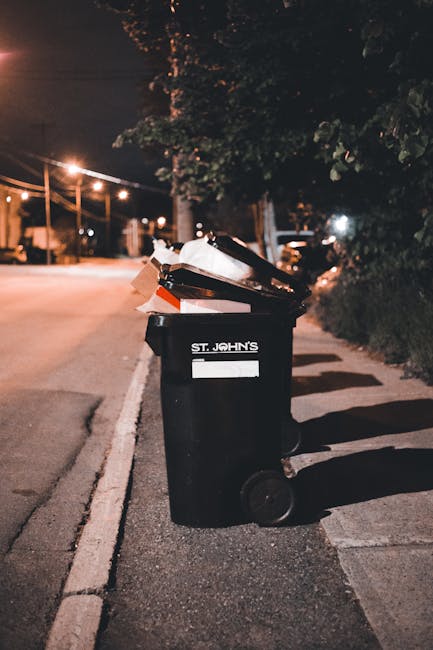 A black wheeled rubbish bin positioned on a pavement beside a street at night, with the label 'ST. JOHN'S' visible on the front. The bin is overflowing with various types of waste, including cardboard boxes, paper, and plastic items, some of which are partially sticking out of the lid. The scene is illuminated by nearby streetlights casting a warm glow, with a background showing trees, parked cars, and distant streetlights along the road. The environment suggests an urban or suburban area where private waste collection services like those offered by Waste Collection Barnet may be used for rubbish removal or on-site waste clearance. The bin's placement on the curb indicates it is awaiting collection, highlighting typical methods of rubbish disposal in residential neighborhoods after private or independent waste management efforts.