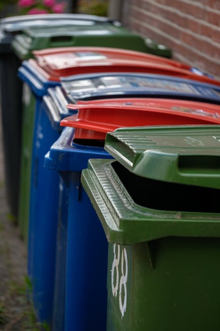 An outdoor scene showing a large collection of discarded cardboard boxes stacked in open-top crates, predominantly featuring fruit shipping cartons with bright, colorful printing and visible labels like 'fresh fruits.' To the right, there are metal wire cages filled with various packed boxes, some topped with brown paper or cardboard lids. In the foreground, several closed plastic waste bins in green, red, and dark colors are visible, with a small shopping cart containing miscellaneous items in front of them. The background includes a paved surface with a faint outline of a garden or rural area, with trees and houses under a partly cloudy sky. The scene reflects an area used for waste collection or disposal, typical of private rubbish removal services, highlighting the organized but cluttered nature of waste handling in an outdoor setting, aligned with the context of alternative waste management options like independent collection or on-site clearance supported by companies such as Waste Collection Barnet.