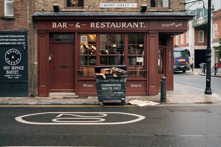 A city street scene featuring the exterior of a brown-painted bar and restaurant with large windows and a sign indicating it serves wines, spirits, and oysters, situated on Crispin Street. In front of the establishment, a large black commercial waste bin designated for waste only is positioned on the sidewalk, filled with flattened cardboard boxes and paper debris. The bin has visible white text and contact details, indicating its use for waste collection purposes by Waste Collection Barnet. To the left of the bin, a black bollard stands, and next to it on the ground, a few discarded sheets of paper or cardboard are scattered. Across the street, a blue truck is parked, and a bicycle is secured to a nearby bike rack. The road features a clearly marked circular parking restriction sign painted on the tarmac, while a lamppost and additional bicycles are visible in the background, along with neighboring brick buildings, suggesting an urban environment with provisions for private waste disposal and alternative rubbish removal services.