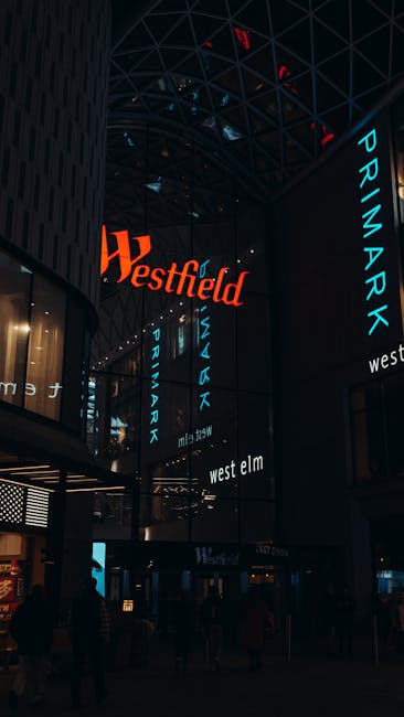 Inside a modern shopping mall with a high glass ceiling, illuminated by artificial lighting, large digital signage displays show the words 'Westfield' and 'PRIMARK' in bright red and blue text respectively, with the signs hanging vertically along the dark metallic framework. The scene includes a spacious, multi-level interior with visible storefronts and walkways on the lower level. In the foreground, a few silhouettes of shoppers and visitors can be seen walking or standing, emphasizing the busy retail environment. Although the image primarily highlights the commercial and architectural aspects of the mall, it subtly relates to waste management services by depicting a setting where private waste collection or on-site clearance might be necessary for maintaining cleanliness in such large retail spaces, aligning with services provided by waste collection companies like Waste Collection Barnet.