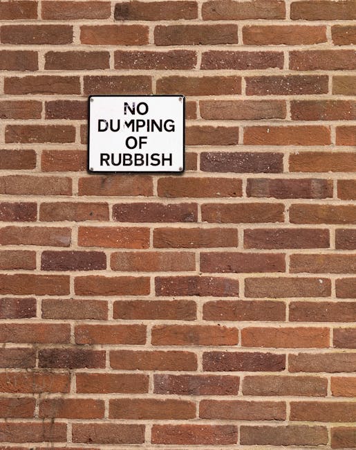 A rectangular white metal sign with black uppercase text reading 'NO DUMPING OF RUBBISH' is mounted on a red brick wall. The brick wall features evenly laid bricks in shades of reddish-brown and dark brown, with light grey mortar joints forming a consistent pattern. The sign is positioned in the upper left quadrant of the image, centered horizontally but towards the top vertically. The wall's surface has a rough texture typical of standard brickwork, and the lighting appears natural, casting minimal shadows. The scene suggests an exterior environment, possibly in a residential or commercial area where waste collection or rubbish removal services, such as those provided by Waste Collection Barnet, are applicable to private or local waste management. The image's focus on the sign and brick wall highlights the importance of designated areas for rubbish disposal and the enforcement of waste removal regulations.