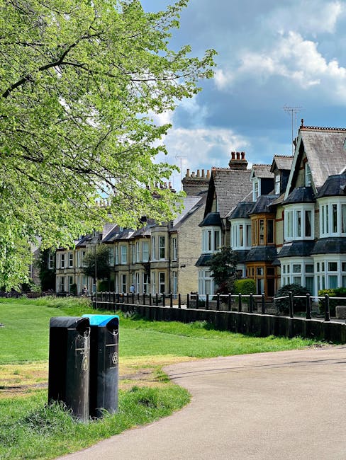 A park area adjacent to a residential street features two black rubbish bins positioned on a grassy patch near a paved footpath. The bins have a simple, functional design with rounded tops and are marked with small white labels. To the left, a large deciduous tree with bright green leaves extends its branches across the upper portion of the image, providing partial shade and adding natural greenery to the scene. In the background, a row of Victorian-style terraced houses with light-colored brickwork, bay windows, and slate roofs line the street, each with small front gardens separated by low wooden fences. The sky above is partly cloudy with patches of blue, creating a typical indeterminate weather day. The overall scene suggests an urban residential area where private waste collection or on-site rubbish disposal methods like bin storage are common, supporting the context of efficient waste management separate from standard municipality collection services, as managed by wastecollectionbarnet.org.uk.